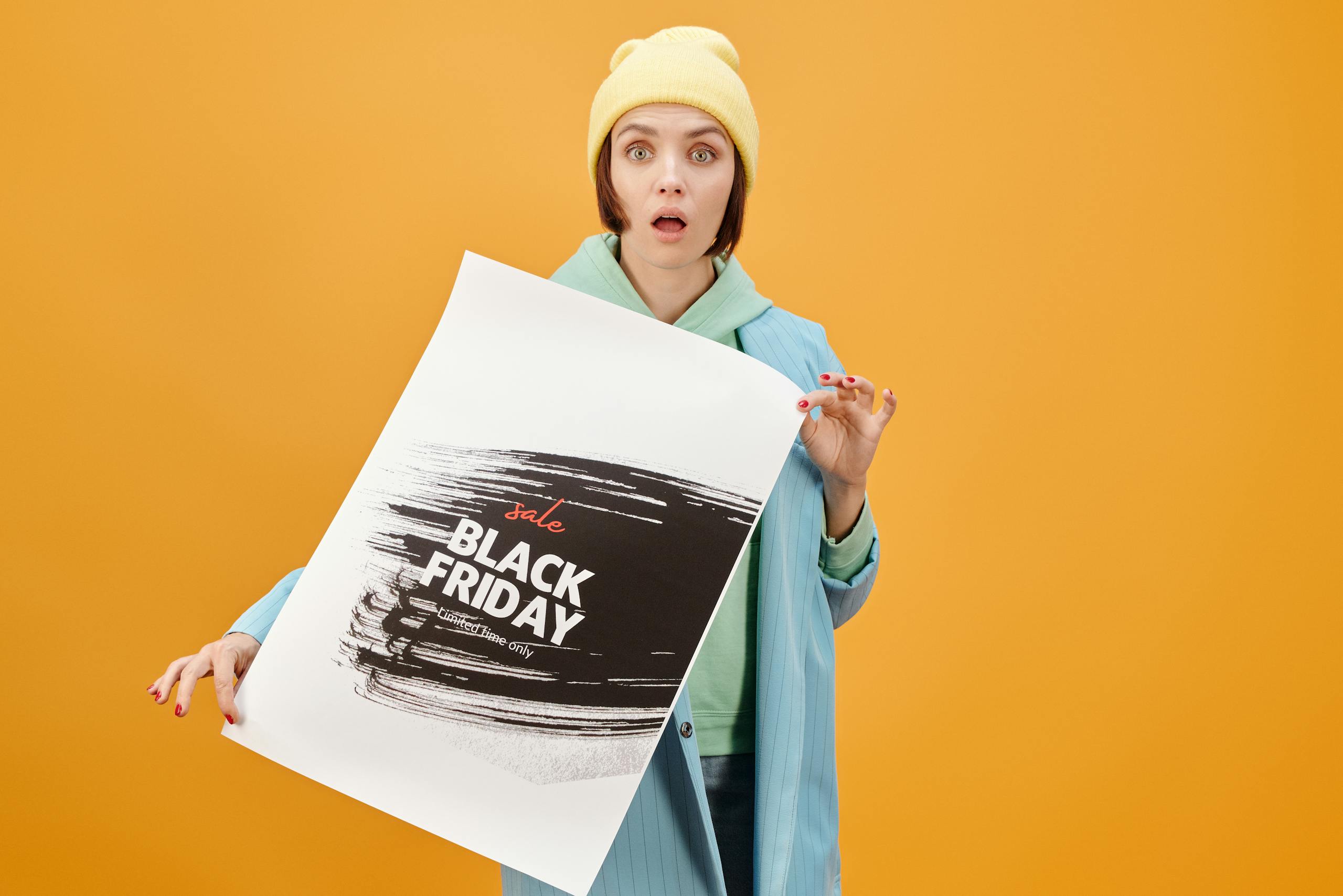 Young woman holds a Black Friday sale poster, surprised against a yellow background.